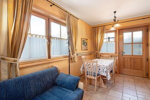 Una habitación con sofá azul, una mesa de comedor de madera, sillas, una ventana con cortinas, una puerta y una lámpara fijada a la pared.