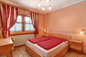 Una habitación con cama, escritorio, silla, ventana con cortinas, radiador e iluminación en la pared.