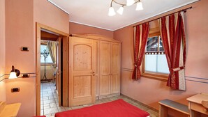 A bedroom with a wooden wardrobe, a bed with red bedding, a desk, and a window with curtains. - Chalet Betty Apartment Muf with Mountain View, Wi-Fi, Balcony & Garden (Livigno)