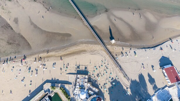 Luchtbeeld van een strand met een lange steiger, een zwembadgedeelte en strandstoelen.