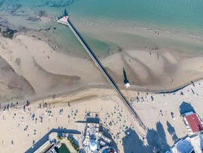 Vista aérea de una playa con un muelle largo, un área de piscina y sillas de playa.