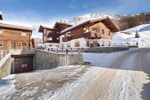 A wooden chalet with a stone entrance, a balcony, and a view of a snowy mountain.