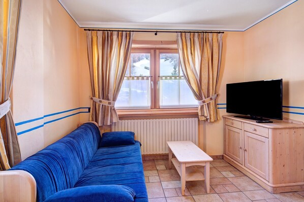 A room with a blue sofa, a wooden table, a TV on a cabinet, and a window with curtains.