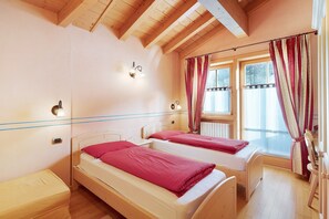 A room with two beds, wooden beams on the ceiling, a window with curtains, and wall-mounted lights.
