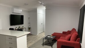 A modern living room with a white sofa, a red sofa, a small table, a TV mounted on the wall, a ceiling fan, and a white cabinet with drawers. - YARRAWONGA PARK COTTAGE<br>Cozy 2 bedroom cottage on small acreage  (Mudgee)