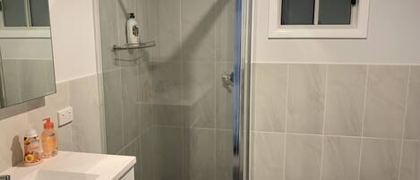 Een badkamer met een douche met glazen wand, een witte wastafel met spiegel en een raam boven de wastafel.