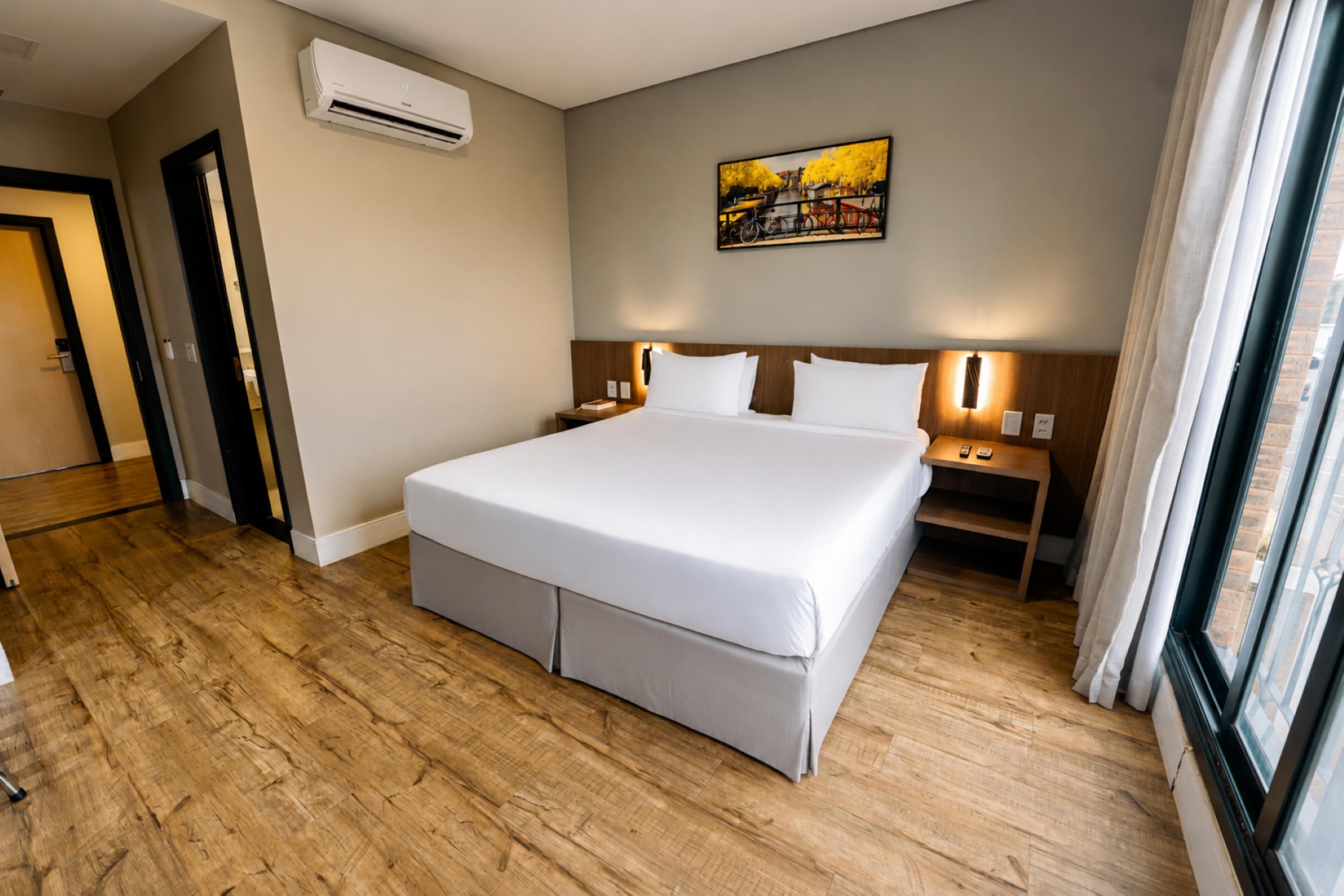Habitación de hotel con una cama grande, piso de madera, unidad de aire acondicionado, mesita de noche, lámpara y ventana con cortinas.