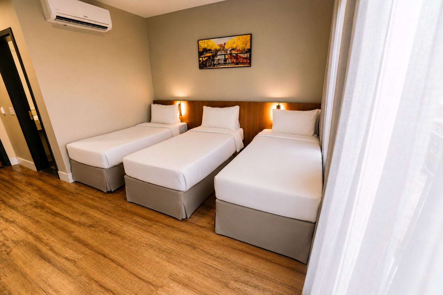 A hotel room with three single beds, a wooden floor, a painting on the wall, and an air conditioning unit.