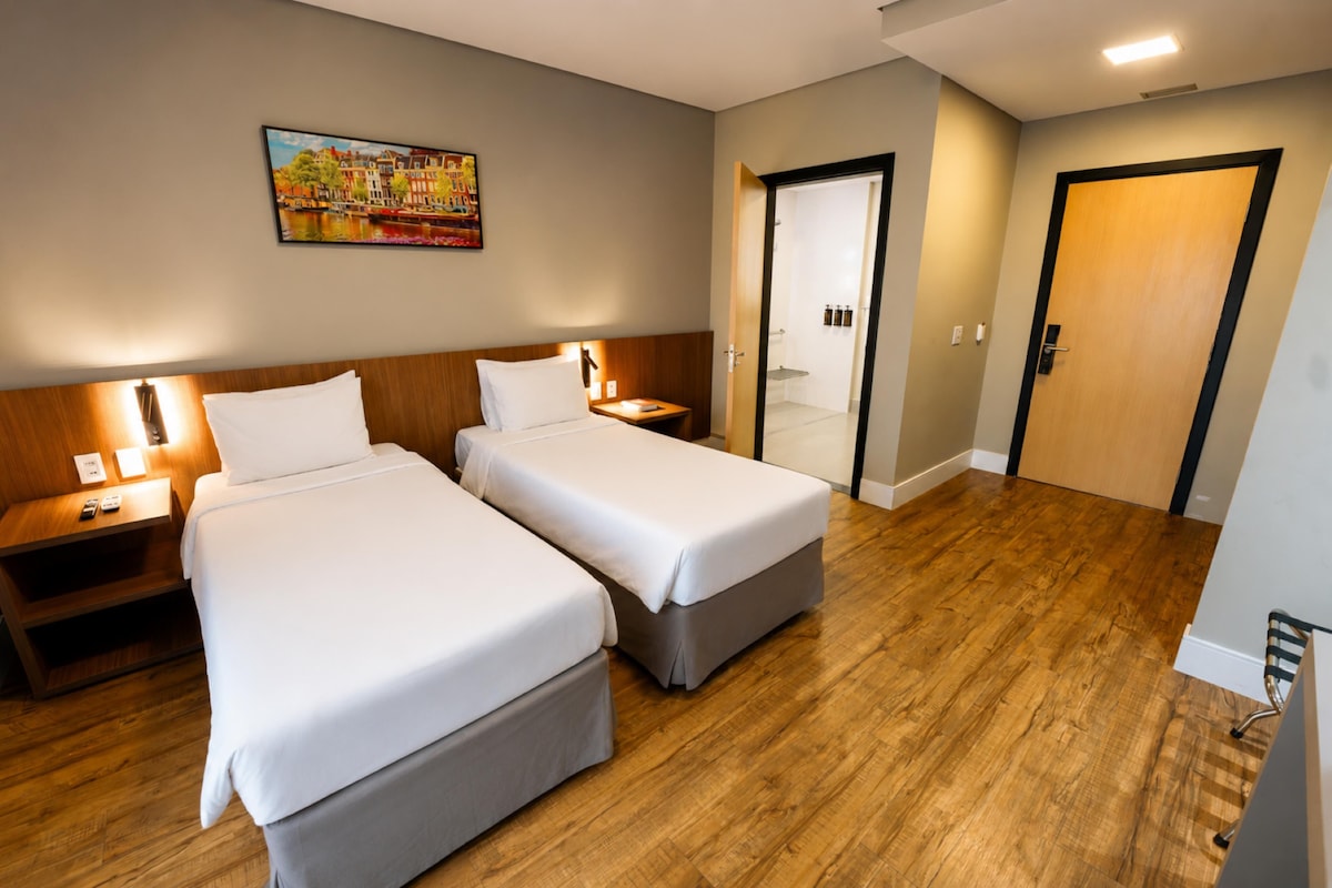 Habitación de hotel con dos camas, piso de madera, un cuadro en la pared y una puerta que da a un baño.