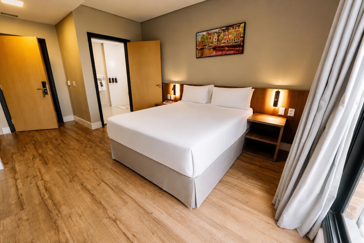 Una habitación de hotel con una cama grande, piso de madera, una puerta, una mesita de noche con una lámpara y un cuadro en la pared.