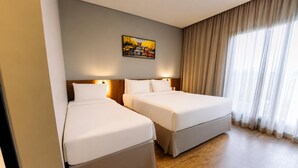 A hotel room with two beds, a wooden floor, a painting on the wall, and a window with curtains.