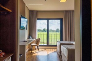 A hotel room with a desk, chair, two beds, and a balcony with a view of the greenery.
