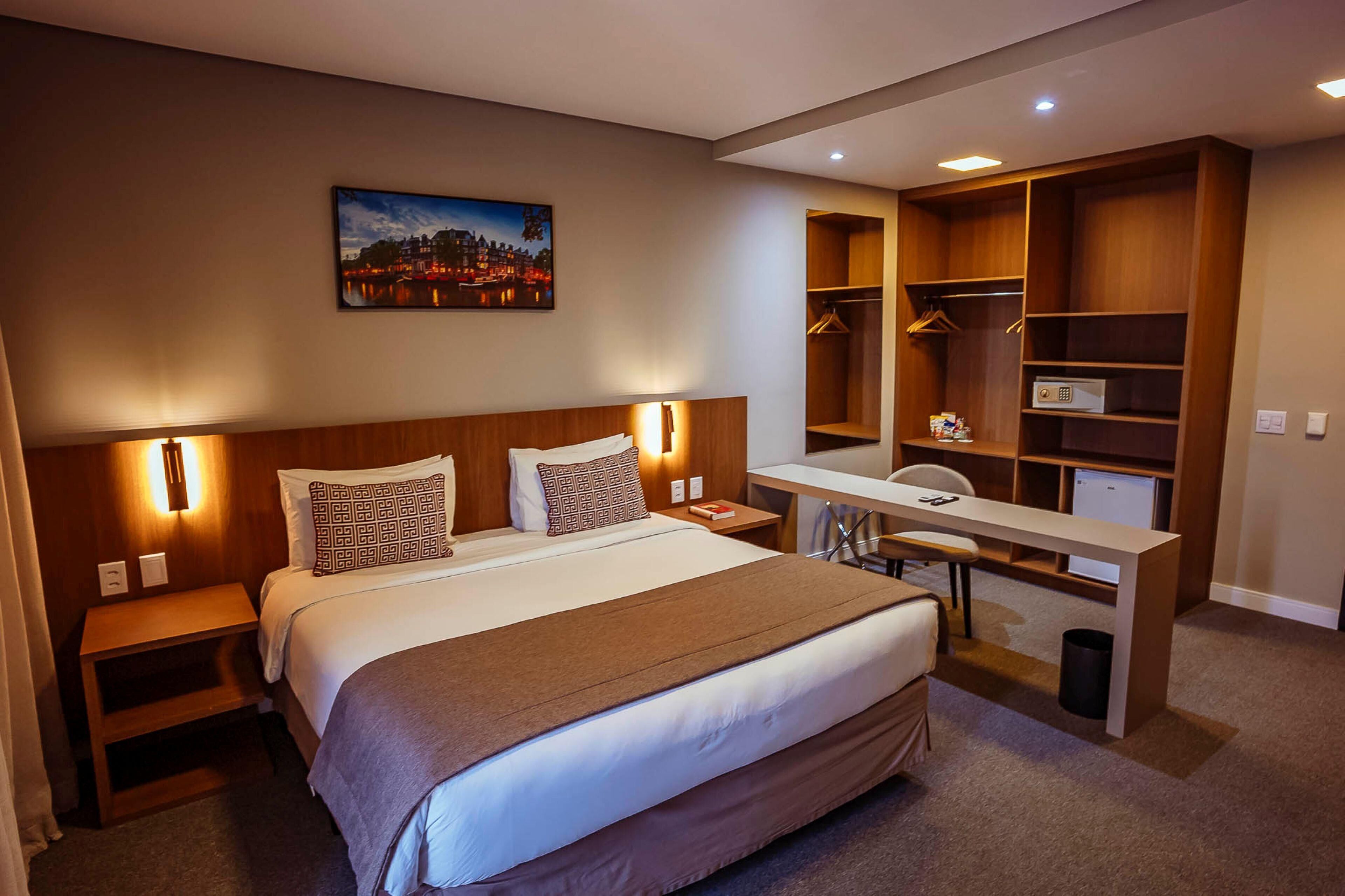 A hotel room with a bed, bedside tables, a desk, a chair, a wardrobe, and a cityscape painting on the wall.