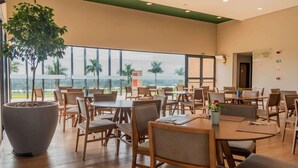 A spacious indoor dining area with multiple tables and chairs, large windows offering a view of palm trees and greenery, and a large potted plant in the foreground.