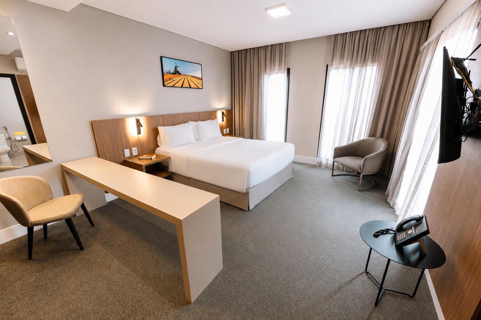 Une chambre d’hôtel avec un lit, un bureau, une chaise, une petite table, un téléviseur et un tableau au mur.