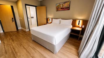 A hotel room with a large bed, wooden flooring, a door, a nightstand with a lamp, and a painting on the wall.