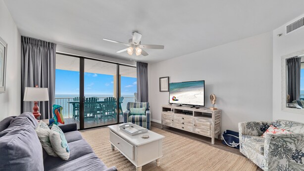 A modern living room with a large TV, a ceiling fan, a sofa, a coffee table, and a view of the ocean through large windows. - Summer House 1403B- Family friendly complex to enjoy the Beach!!! (Orange Beach)