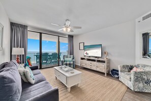 A modern living room with a large TV, a ceiling fan, a sofa, a coffee table, and a view of the ocean through large windows. - Summer House 1403B- Family friendly complex to enjoy the Beach!!! (Orange Beach)