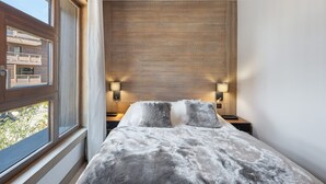 A bedroom with a large window, wooden panel wall, two bedside lamps, and a fluffy grey bedspread. - Wellness Getaway: Modern Apartment in Megève with Pool, Gym, and Spa Access (Megève)