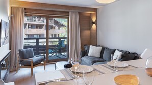 A modern hotel room with a dining area, a sofa, and a balcony with wooden railings. - Wellness Getaway: Modern Apartment in Megève with Pool, Gym, and Spa Access (Megève)