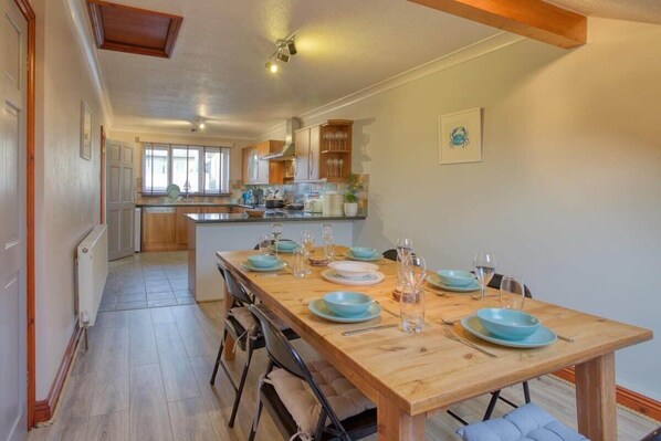 A dining area with a wooden table set for four, a kitchen in the background, and a framed picture on the wall.