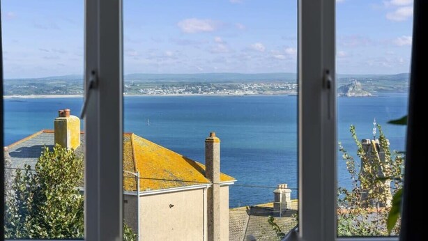 A coastal view from a window, including a building with a yellow roof and chimneys, and a distant shoreline with buildings and a clear sky. - Large House with Panoramic Sea Views & Parking (Newlyn)