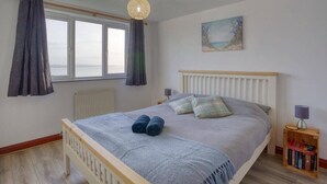 A bedroom with a bed, curtains, a window, a radiator, a bedside table with a lamp, and a painting on the wall. - Large House with Panoramic Sea Views & Parking (Newlyn)
