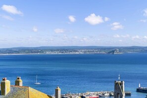 A coastal town with a prominent stone tower, a harbor with boats, and a clear view of the sea and distant land.