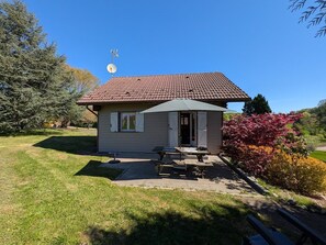 Una pequeña casa con techo de tejas, una puerta y una ventana con contraventanas. Hay una mesa de picnic y un banco en el jardín.