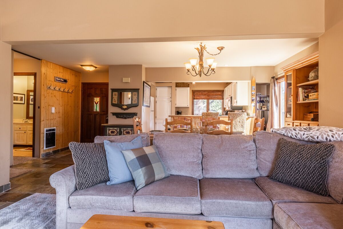 A living room with a grey sectional sofa, wooden flooring, a chandelier, and a kitchen in the background.