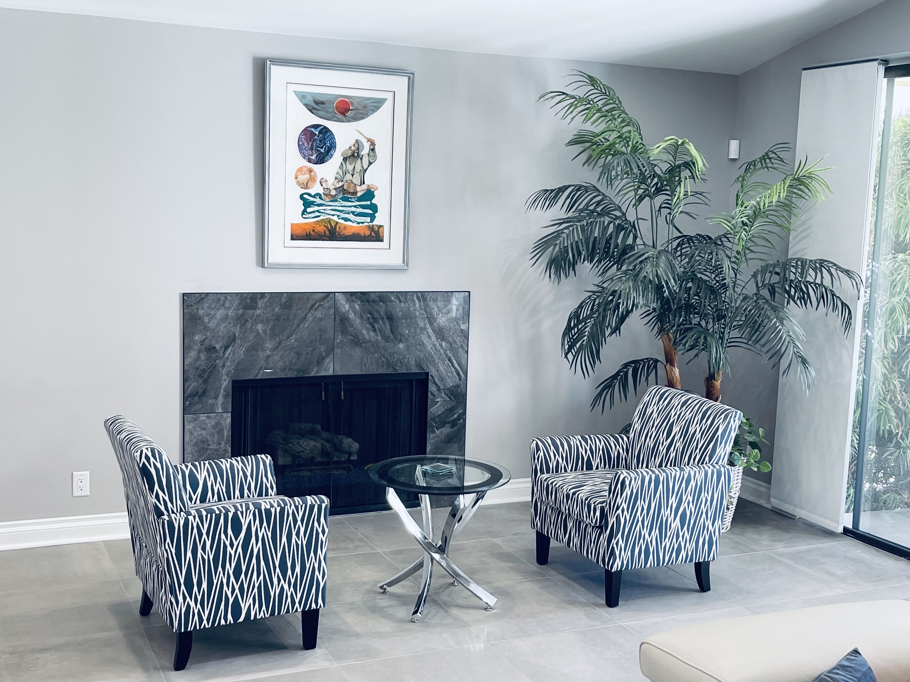 A modern living room with a fireplace, two patterned armchairs, a glass coffee table, and a large potted plant.