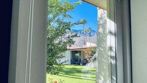 Uitzicht door een raam op een huis met een terras, een gazon en bergen op de achtergrond.