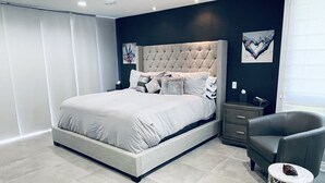 Een moderne slaapkamer met een bed met gestuikt hoofdbord, een grijze fauteuil, een nachtkastje en een decoratief wandobject.