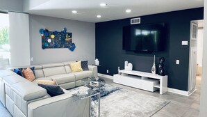 Een moderne woonkamer met een hoekbank, een glazen salontafel, een flatscreen televisie aan de muur en een decoratief kunstwerk.