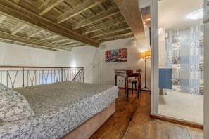 Une chambre avec un plafond en bois, un lit recouvert d’une couvre-lit à motifs floraux, un bureau avec une chaise et un tableau accroché au mur.