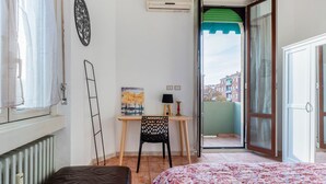 Ein Schlafzimmer mit einem Bett, einem Schreibtisch mit Stuhl, einem Heizkörper, einem Fenster mit Vorhängen und einer offenen Tür, die auf einen Balkon führt.