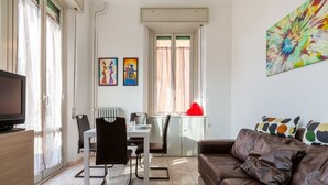 Ein modernes Wohnzimmer mit einem braunen Ledersofa, einem Essbereich mit weißem Tisch und Stühlen und einem Fernseher links.