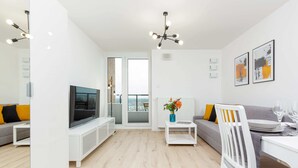 A modern living room with a flat-screen TV, a sofa with yellow cushions, a dining table with a white chair, and artwork on the wall.