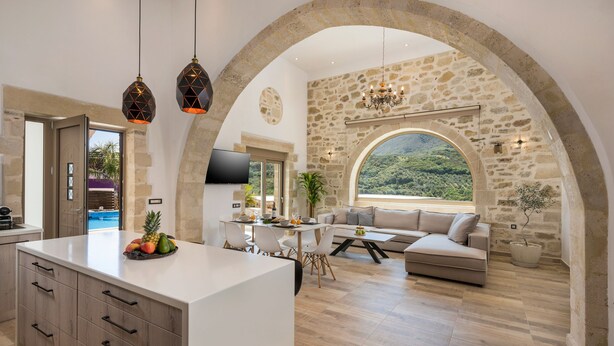 A spacious living area with a stone wall, a large window, and a modern kitchen. - Casa Belvedere Boutique Villa (Apokoronas)