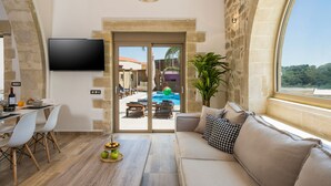 A modern living room with a sofa, dining table, and chairs, a TV mounted on the wall, and a view of an outdoor pool area. - Casa Belvedere Boutique Villa (Apokoronas)