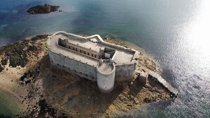 An aerial view of a coastal fortification with a central tower, surrounded by a moat and a sandy beach.