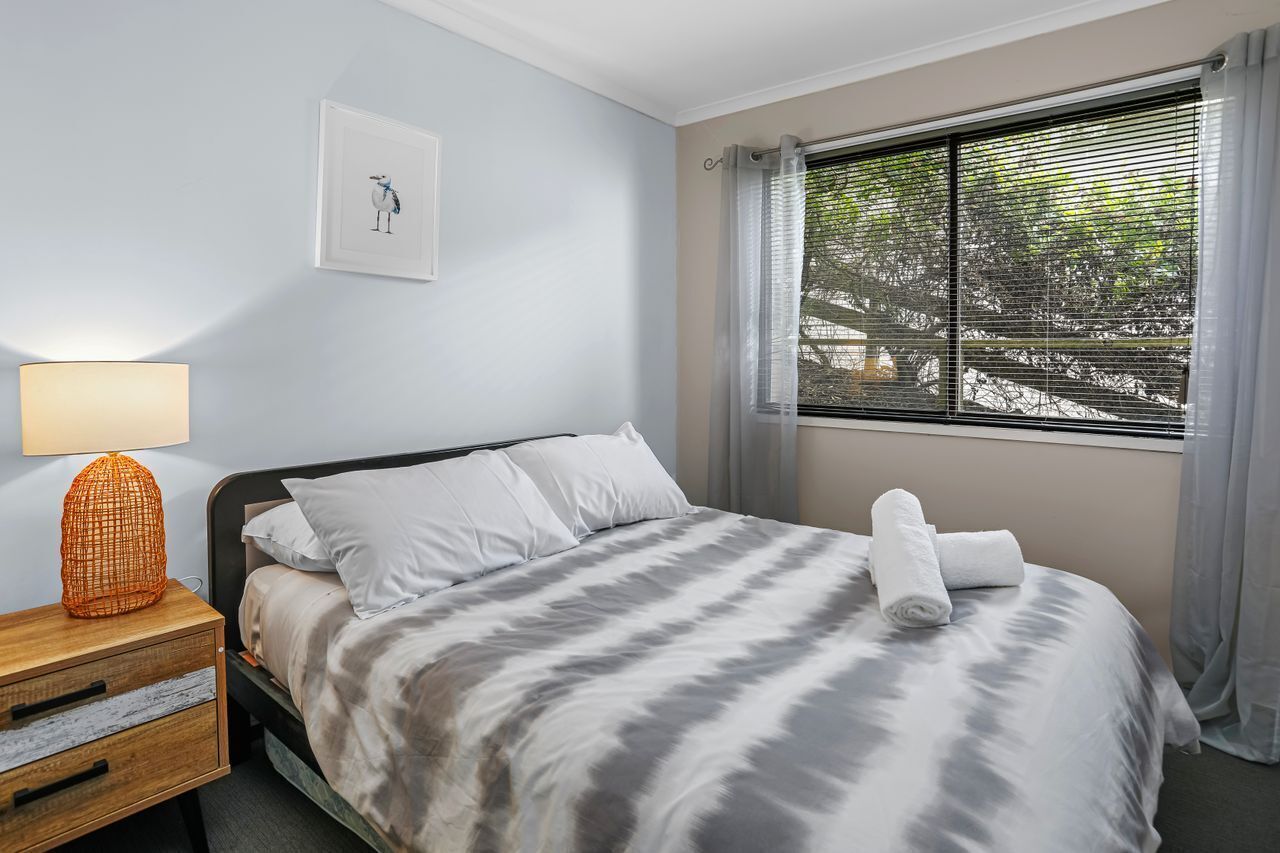 A neatly made bed with a striped bedspread, a bedside lamp, a framed picture on the wall, and a window with curtains.