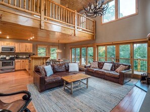 A spacious living area with a high ceiling, wooden flooring, and large windows offering a view of the outdoors.