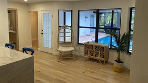 Quarto com piso de madeira, uma cadeira de vime, porta branca e grandes janelas com vista para uma piscina.