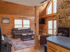 Interior de una cabaña de madera con sala de estar, cocina, pared de piedra y un televisor montado en la pared.