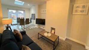 A modern living room with a sofa, coffee table, and TV. There is a dining area with chairs and a table. The room has a large window and a ceiling fan. - *Exclusive* Modern, Stylish 3-bed, sleeps 6 -Birmingham,Solihull,NEC,Airport,HS2 (Birmingham)