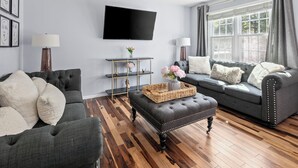 A living room with a grey sofa, a matching ottoman, and a flat-screen TV mounted on the wall. - GannonHeights: Work-Friendly & 40 mins to DC (Sterling)