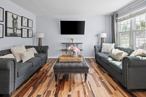 A living room with two grey sofas, a central ottoman, a flat-screen TV, and framed artwork on the wall.