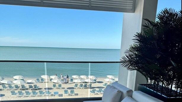 Ein Balkon mit einer weißen Couch und runden Tischen, mit Blick auf einen Strand mit Sonnenschirmen und Liegestühlen.
