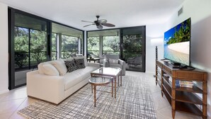 Ein modernes Wohnzimmer mit einem weißen Sofa, einem Glastisch und einem Fernseher auf einem Holzständer. Große Fenster bieten Blick ins Grüne.